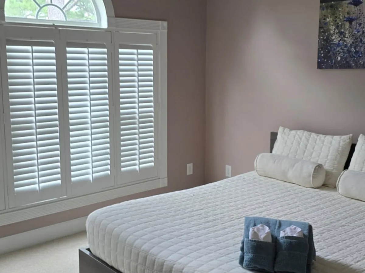 Organized bedroom with white bedding, folded towels on the bed and plantation shutters